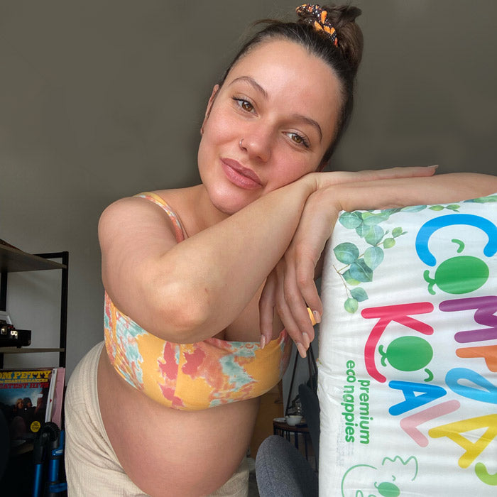Pregnant woman smiling and leaning on a pack of eco-friendly diapers from 'Comfort Koala.'